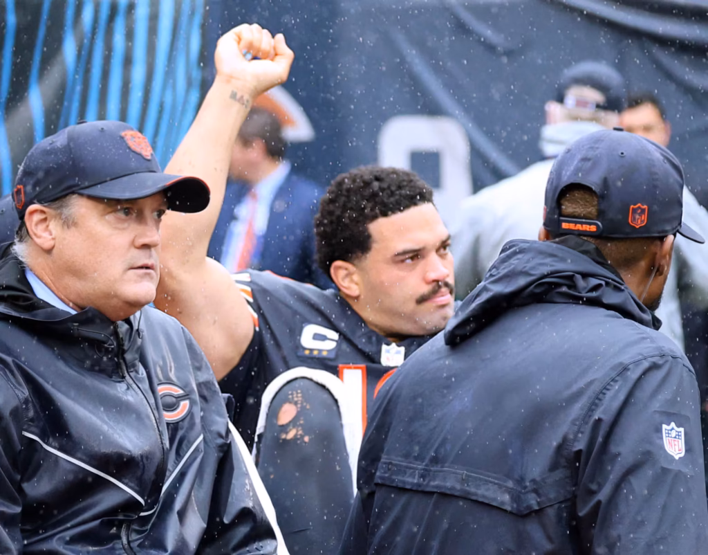 Bears quarterback raises an arm while surrounded by staff in rain.